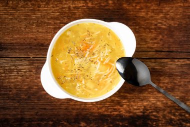 Erişteli tavuk çorbası, patates, soğan ve havuç, ahşap arka planda beyaz bir tabakta..