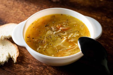 Erişteli tavuk çorbası, patates, soğan ve havuç siyah kaşık ve ahşap arka planda beyaz ekmek ile beyaz bir tabakta..