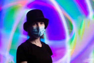 Tıbbi maskeli bir kadın ve siyah şapkalı bir kadın. Arka planda çok renkli bir ışık var..