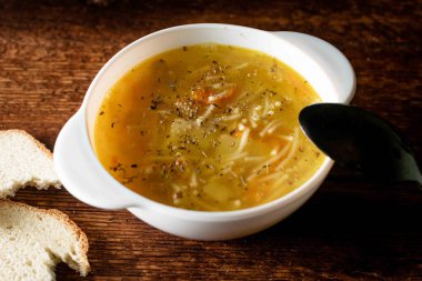 Erişteli tavuk çorbası, patates, soğan ve havuç siyah kaşık ve ahşap arka planda beyaz ekmek ile beyaz bir tabakta..