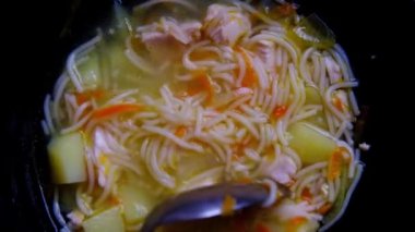 Bir tabak et çorbası, spagetti, patates siyah kaşıkla karıştırılır..