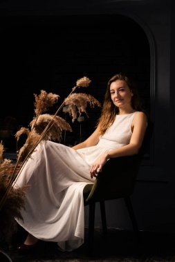 Beyaz elbiseli genç bir kadın düşünceli bir şekilde karanlık bir odada koltukta oturur. Yalnızlık, içgözlem konsepti.