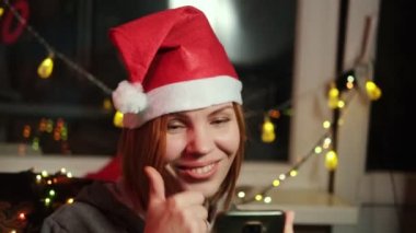 Yeni yıl şapkalı orta yaşlı bir kadın telefonda video görüşmesiyle gülümsüyor ve iletişim kuruyor. Noel 'e kadar yeni yıl tatili için tebrikler..