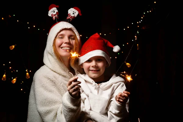 母親と息子は 彼らの手の中に火花で幸せと笑顔です 暗闇の中でクリスマスの帽子の男の子と女性 — ストック写真