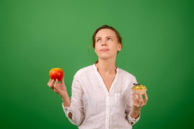 Elmalı ve pastalı orta yaşlı bir kadın kendi seçimiyle eziyet görüyor. Yeşil bir arka planda. Doğru sağlıklı yiyeceği seçme fikri..