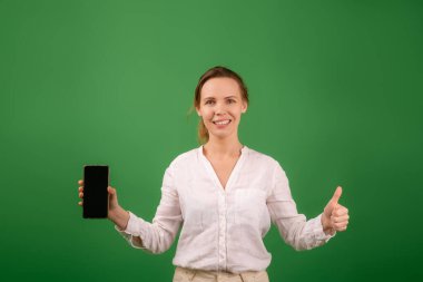 Beyaz tişörtlü çekici orta yaşlı bir kadın gülümsüyor ve elinde yeşil ekranlı bir cep telefonu tutuyor. Model.