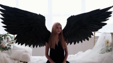Siyah melek kanatları olan bir kız beyaz bir yatakta oturur..