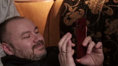 Elinde telefonla gülümseyen orta yaşlı bir adam. Yarı karanlık bir odada, kanepede yatıyor. Sanal iletişim.