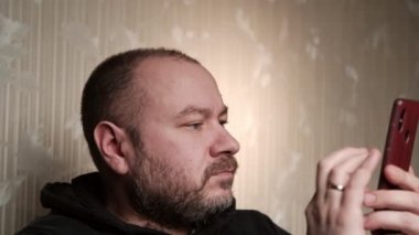 Elinde telefonla gülümseyen orta yaşlı bir adam. Yarı karanlık bir odada, kanepede yatıyor. Sanal iletişim.