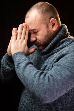 A middle-aged man is praying. Unshaven man in a sweater. Dark background.