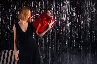 Elinde kırmızı bir hediye olan bir kadın. Parlak bir duvarda parti yazıları. Kırk yaşında, orta yaşlı bir kadın, gece elbisesi giymiş. Neşeli, mutlu yüz. Kutlama, parti konsepti. Hediyeler ver. Metnin için yer.