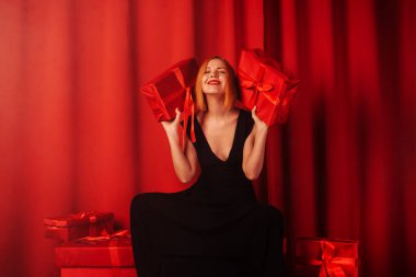 An amazed, joyful woman sits on a multitude of gifts. Middle-aged woman in a black dress. Red background. Happy face. Giving, receiving gifts concept.