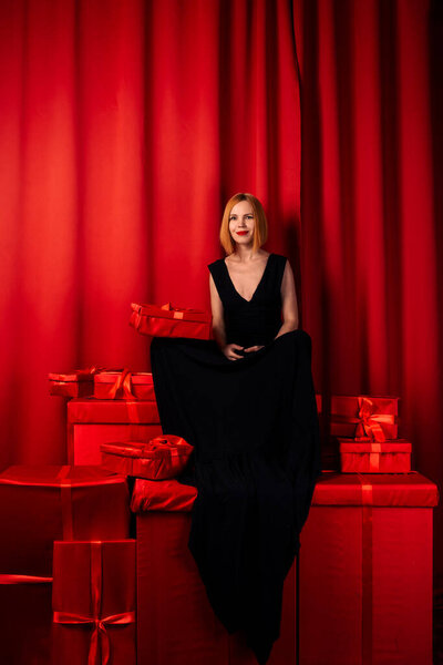 Женщина и много красных подарков. Женщина средних лет сидит на упакованных подарках. Черное длинное платье. Красный фон. Счастливое лицо.