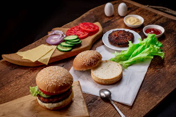 Cooking hamburger or cheeseburger. Different ingredients for a classic hamburger. Grilled meat, vegetables, greens, sauces near a sesame bun. Wooden background. Hamburger day.