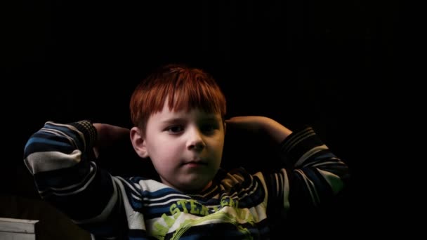 若い赤髪の少年は落ち着いて座って暗い背景にカメラを見て — ストック動画