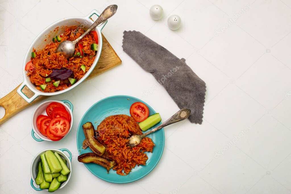 Jollof rice with fried banana on a plate. Rice with tomatoes, onions, spices. Fresh vegetables. White background. View from above.