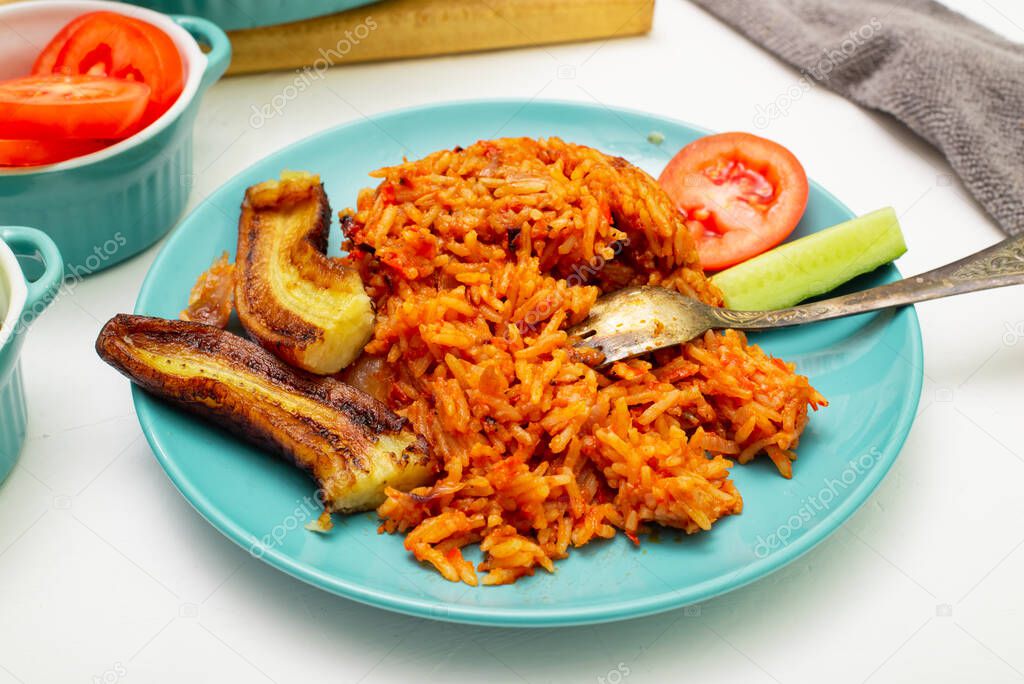 Jollof rice with fried banana on a plate. Rice with tomatoes, onions, spices. Fresh vegetables. White background.