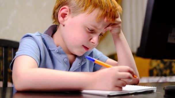 小学生勤勤恳恳 坐在桌旁紧张地思考着 在家上学 一个红头发的小男孩 — 图库视频影像