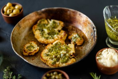 Baharatlı karnabahar bifteği kızartma tavasında yatıyor. Zeytinyağı, chimichurri sosu, kapari, zeytin, otlar, çeşitli baharatlar yan yana. Karanlık arka plan. Vejetaryen yemeği..
