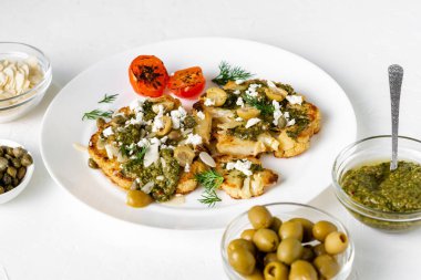 Baharatlı karnabahar bifteği, chimichurri sosu, badem gevreği, zeytin, kızarmış vişneli domates ve beyaz tabakta kapari. Vejetaryen yemeği. Beyaz arkaplan.