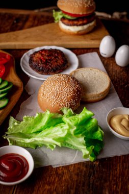 Hamburger ya da çizburger pişirmek. Klasik bir hamburger için farklı malzemeler. Izgara et, sebze, sebze, susamlı ekmeğin yanında soslar. Ahşap arkaplan.