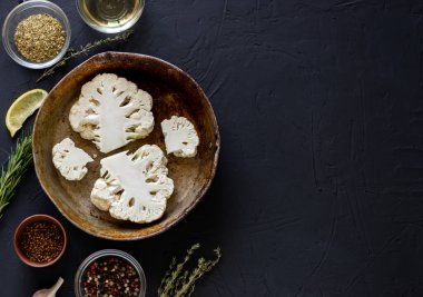 Karnabahar bifteği pişirme. Çiğ kesilmiş karnabahar kızartma tavasında yatıyor. Zeytinyağı, bitkiler, yakınlarda çeşitli baharatlar. Karanlık arka plan. Metin için yer.