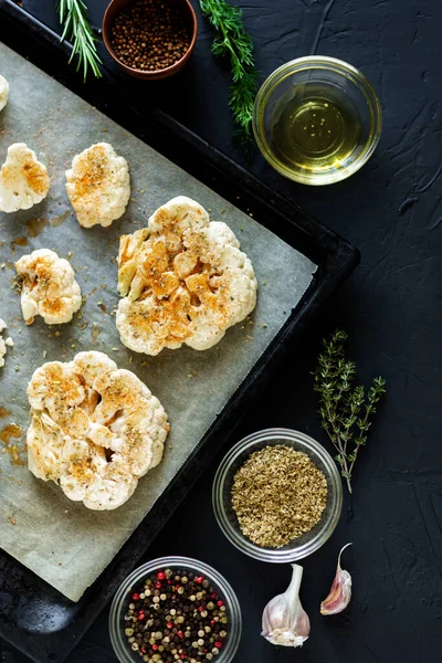 Karnabahar bifteği pişirme. Fırın örtüsünün üzerine baharat serpiştirilmiş çiğ karnabahar. Zeytinyağı, bitkiler, yakınlarda çeşitli baharatlar. Koyu arkaplan.