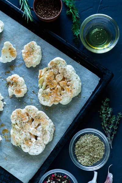 Karnabahar bifteği pişirme. Fırın örtüsünün üzerine baharat serpiştirilmiş çiğ karnabahar. Zeytinyağı, bitkiler, yakınlarda çeşitli baharatlar. Karanlık arka plan. Renkli.