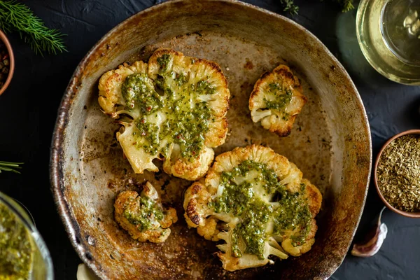 Baharatlı karnabahar bifteği kızartma tavasında yatıyor. Zeytinyağı, chimichurri sosu, otlar, çeşitli baharatlar yan yana. Karanlık arka plan. Vejetaryen yemeği..