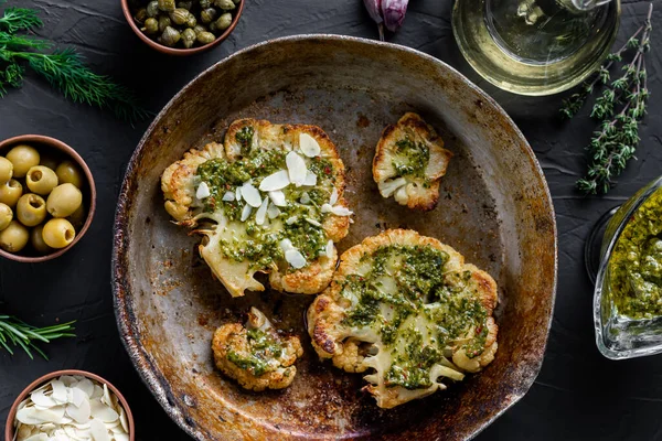 Baharatlı karnabahar bifteği kızartma tavasında yatıyor. Zeytinyağı, chimichurri sosu, kapari, zeytin, otlar, çeşitli baharatlar yan yana. Karanlık arka plan. Vejetaryen yemeği..