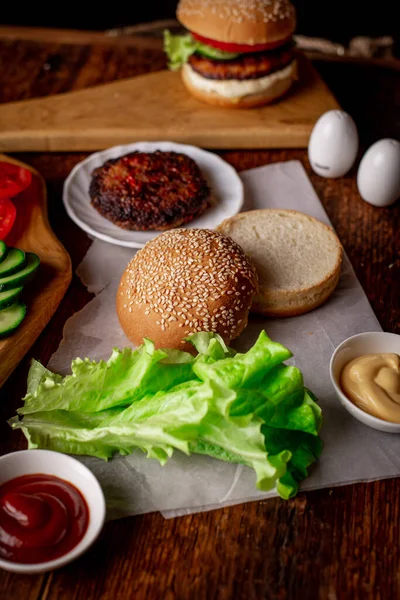Hamburger ya da çizburger pişirmek. Klasik bir hamburger için farklı malzemeler. Izgara et, sebze, sebze, susamlı ekmeğin yanında soslar. Ahşap arkaplan.