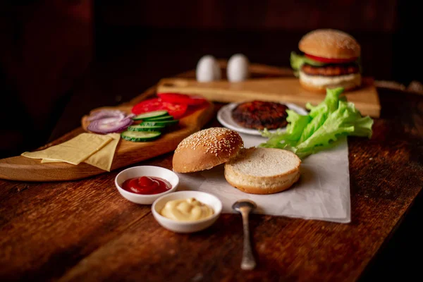 Cooking hamburger or cheeseburger. Different ingredients for a classic hamburger. Grilled meat, vegetables, greens, sauces near a sesame bun. Wooden background. Hamburger day.