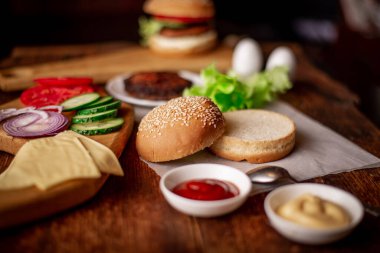 Hamburger ya da çizburger pişirmek. Klasik bir hamburger için farklı malzemeler. Izgara et, sebze, sebze, susamlı ekmeğin yanında soslar. Ahşap arkaplan.
