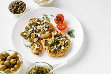 Baharatlı karnabahar bifteği, chimichurri sosu, badem gevreği, zeytin, kızarmış vişneli domates ve beyaz tabakta kapari. Vejetaryen yemeği. Beyaz arkaplan.