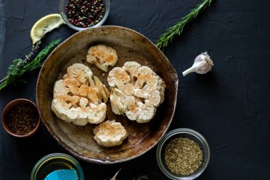 Karnabahar bifteği pişirme. Kızartma tavasına baharat serpiştirilmiş çiğ karnabahar. Zeytinyağı, bitkiler, yakınlarda çeşitli baharatlar. Koyu arkaplan.