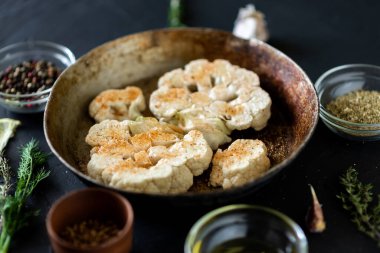 Karnabahar bifteği pişirme. Kızartma tavasına baharat serpiştirilmiş çiğ karnabahar. Zeytinyağı, bitkiler, yakınlarda çeşitli baharatlar. Koyu arkaplan.