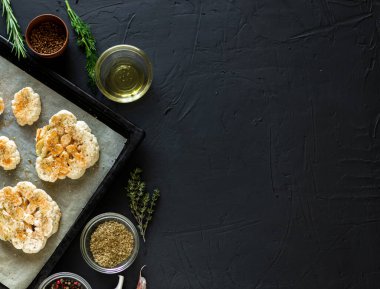 Karnabahar bifteği pişirme. Fırın örtüsünün üzerine baharat serpiştirilmiş çiğ karnabahar. Zeytinyağı, bitkiler, yakınlarda çeşitli baharatlar. Karanlık arka plan. Kopyalama alanı.