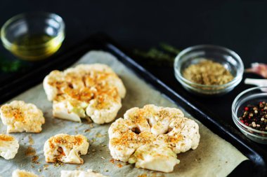 Karnabahar bifteği pişirme. Fırın örtüsünün üzerine baharat serpiştirilmiş çiğ karnabahar. Zeytinyağı, bitkiler, yakınlarda çeşitli baharatlar. Koyu arkaplan.