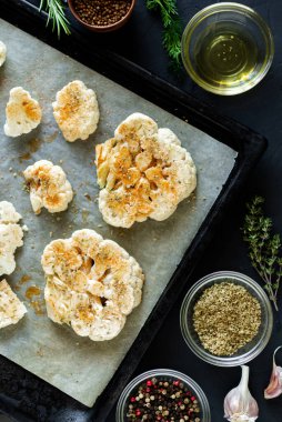 Karnabahar bifteği pişirme. Fırın örtüsünün üzerine baharat serpiştirilmiş çiğ karnabahar. Zeytinyağı, bitkiler, yakınlarda çeşitli baharatlar. Koyu arkaplan.