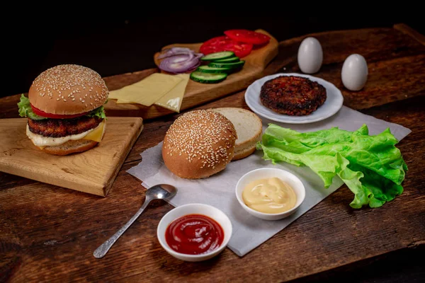 Cooking hamburger or cheeseburger. Different ingredients for a classic hamburger. Grilled meat, vegetables, greens, sauces near a sesame bun. Wooden background. Hamburger day.