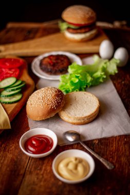 Hamburger ya da çizburger pişirmek. Klasik bir hamburger için farklı malzemeler. Izgara et, sebze, sebze, susamlı ekmeğin yanında soslar. Ahşap arkaplan.