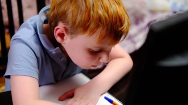 Genç kızıl saçlı bir çocuk ev ödevi yapıyor, uzaktan çalışıyor, bilgisayar ekranının önünde deftere yazıyor. Evde eğitim. Karantina.