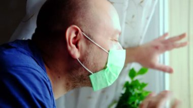 Tıbbi maskeli bir adam perdeyi çeker ve pencereden el sallar. Kişisel izolasyon, hastalık koronavirüs konsepti. Pencereden tarih.