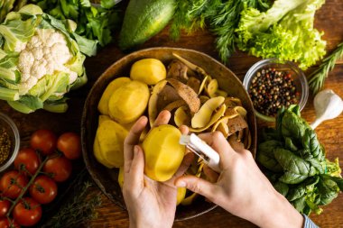 Bir kadın patates soyuyor. Masanın üzerinde çeşitli taze sebzeler var. Vejetaryen yemeği pişirmek.