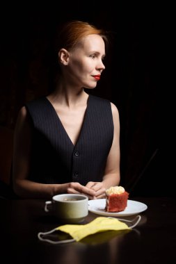 Masada elinde çay ve kek olan bir kadın var. Siyah yelek giyen 40 yaşında çekici bir kadın. Çay içiyorum. Masada tıbbi maske.