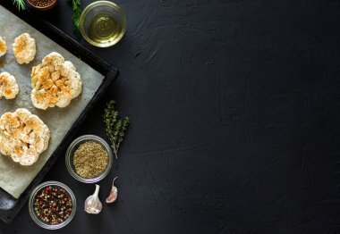 Karnabahar bifteği pişirme. Fırın örtüsünün üzerine baharat serpiştirilmiş çiğ karnabahar. Zeytinyağı, bitkiler, yakınlarda çeşitli baharatlar. Karanlık arka plan. Kopyalama alanı.