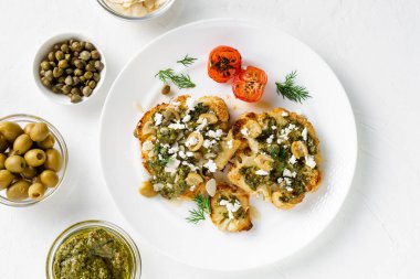 Vejetaryen gıda konsepti. Baharatlı karnabahar bifteği, chimichurri sosu, badem gevreği, zeytin, kızarmış vişneli domates ve beyaz tabakta kapari. Beyaz arkaplan.