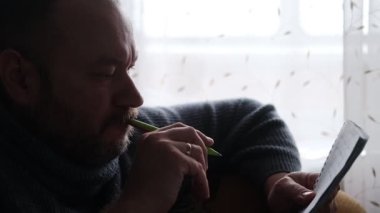 Orta yaşlı bir adam deftere kalemle yazar. Yaratıcılık, yazma. Süveter giyen sakallı bir adam..