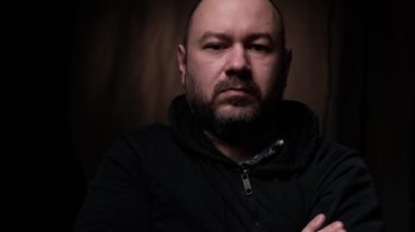 Orta yaşlı bir adam kameraya tarafsız bakıyor. Koyu arkaplan.