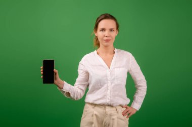 Beyaz tişörtlü çekici orta yaşlı bir kadın gülümsüyor ve elinde yeşil ekranlı bir cep telefonu tutuyor. Model.